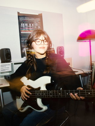 Ung kvinna med glasögon och flätor spelar på en elbas i ett musikstudio-liknande rum med högtalare och musikutrustning i bakgrunden.