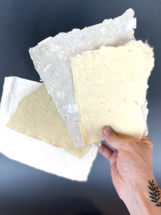 Hand med svart bladtatuer­ing håller tre ark handgjort papper i olika färger.