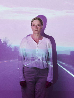 Kvinna i 50-årsåldern står framför en projektion av en landsväg, bilden visas över hennes kropp.