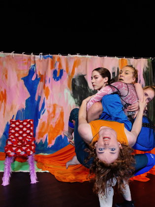 Fyra kvinnor håller i varandra framför en svart scen med en färgglad fond och ett bord med orange duk.