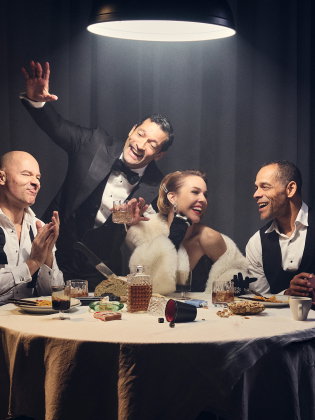 Fyra vuxna personer i festkläder runt ett bord med mat och dryck, en man står upp och gestikulerar.