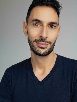 Man i 30–40-årsåldern med kort lockigt hår och skägg, i mörkblå t-shirt mot ljus bakgrund.