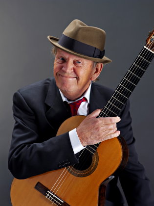 Mikael Neumann leendes iklädd kostym och hatt med en gitarr i handen.