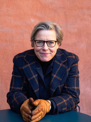 Kvinna i 50-årsåldern med kort blont hår, glasögon och rutmönstrad kappa vid orange vägg.