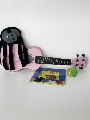 Rosa ukulele med en svart spindelgosedjur ovanpå, barnbok och grön snigelleksak framför.