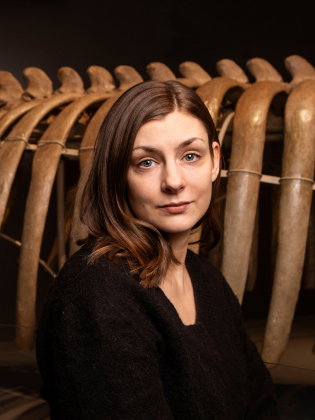 Kvinna i 30-årsåldern med brunt axellångt hår och svart tröja, står framför valskelett.