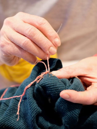 En person i grå T-shirt lagar ett mörkgrönt plagg med rosa garn och nål.