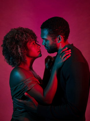 En kvinna och en man i 30-årsåldern står tätt, tittar varandra i ögonen under färgat ljus.