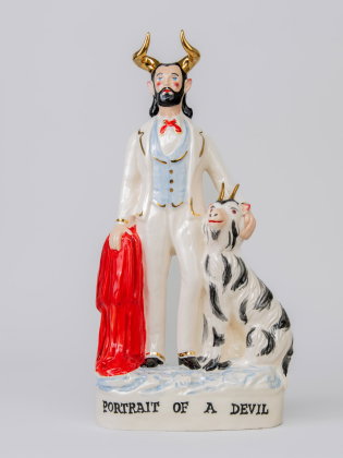 Porslinsfigur av en man med horn, skägg och kostym, hållande en röd mantel bredvid en getliknande varelse med horn.