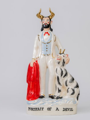 Porslinsfigur av en man med horn, skägg och kostym, hållande en röd mantel bredvid en getliknande varelse med horn.