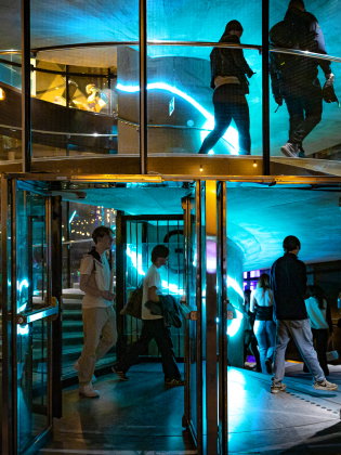 Flera personer går in genom glasad karuselldörr till en byggnad med neonljus.