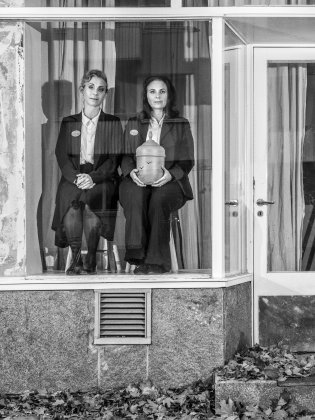 Helen Sjöholm och Anna Fahlstedt sitter på varsin pall i ett tomt skyltfönster. I sina händer har Anna en begravningsurna.