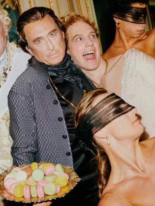 Fem personer i barockinspirerade kläder, två med ögonbindlar, en håller ett fat med färgglada macrons.