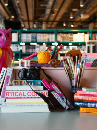 Bord fullt av färgglada konstmaterial, böcker, penslar, kamera och en rosa uggla i ett bibliotek.