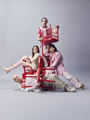 Fyra vuxna, två män och två kvinnor, poserar med staplade röda stolar mot grå bakgrund i rosa och beige kläder. En man ligger under.