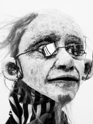 Närbild av docklik äldre kvinna med rynkigt ansikte, runda glasögon av metalltråd och grått hår mot vit bakgrund.