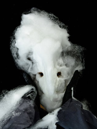 Person med vitt, fibermaterial format som mask över ansiktet, hålls på plats av svarta handskar mot svart bakgrund.