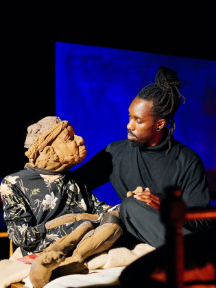 Vuxen man i svarta kläder sitter på säng och håller en docklik äldre kvinna i blommig klänning på en teaterscen.