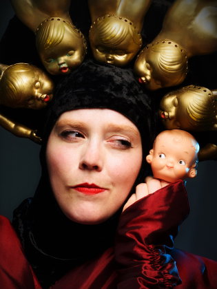Kvinna i 30–40-årsåldern med rött läppstift och svart huvudbonad med guldfärgade dockhuvuden, håller liten babydocka.