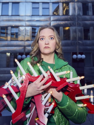 Kvinna i grön jacka står utomhus framför en byggnad och håller flera röda adventsljusstakar med tända lampor i famnen.