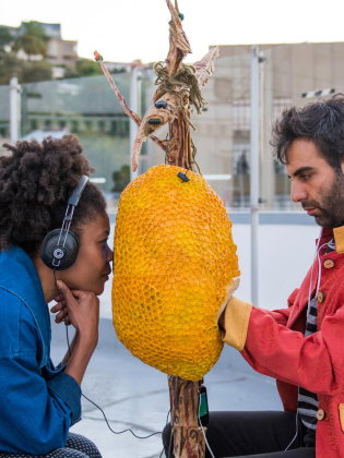 Två personer sitter mittemot varandra utomhus och lyssnar via hörlurar på ett stort gult objekt upphängt mellan dem.