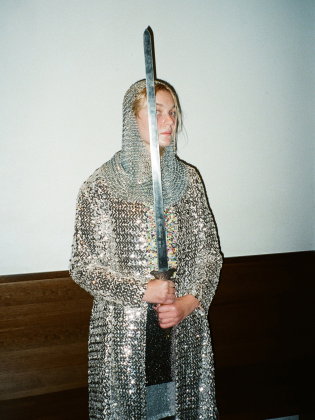 Person i glittrande rustning och ringbrynja står med ett stort svärd framför kroppen och ser åt sidan mot en neutral vägg.