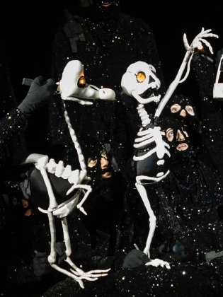 Flera vuxna i svarta, glittriga dräkter manipulerar skelettdockor med lysande ögon på mörk scen.