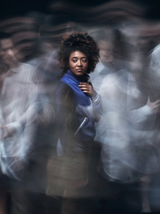 Kvinna ca 25–35 med afro och blå jacka står stilla med hand på bröstet medan suddiga personer rör sig runt henne.