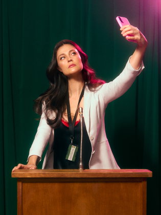 Kvinna vid en talarstol tar en selfie med mobilen, med namnbricka runt halsen och grön ridå i bakgrunden.