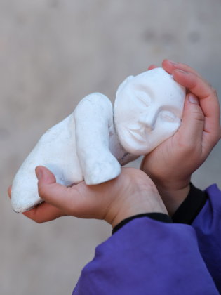 Ett person håller en liten vit skulptur av en liggande figur med slutna ögon i sina händer. Närbild mot en mjuk, neutral bakgrund.