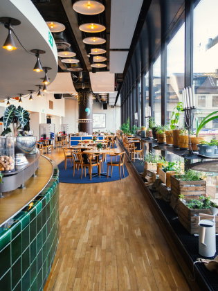 Ljus restaurang med trägolv, runda bord och stolar. Stora fönster längs väggen med krukväxter och utsikt över stadsgata.