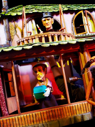 Interiör av modellhus med flera dockfigurer; en håller en låda medan en hundlik figur står i förgrunden.