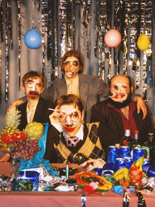 Fyra personer sitter vid ett festbord fullt av frukt, burkar och flaskor. De bär tejpade ansiktsbilder som masker mot glittrig bakgrund.