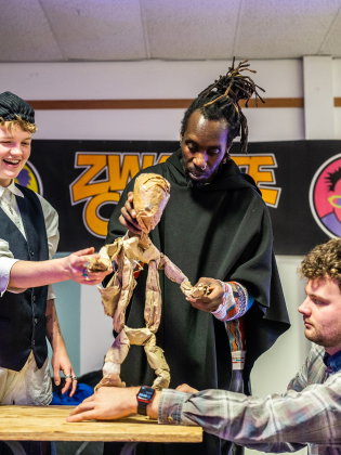 Tre personer samarbetar kring en docka av papper i ett rum, de skrattar och fokuserar under en workshop eller repetition.