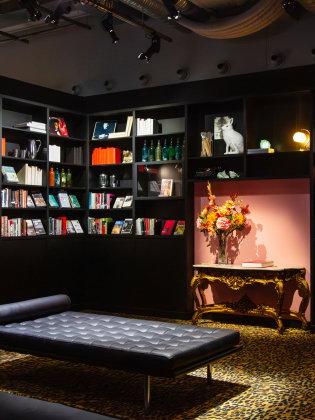 Stilfullt rum med svarta bokhyllor fyllda med böcker och objekt, en låg schäslong och ett bord med blomvas i varm belysning.