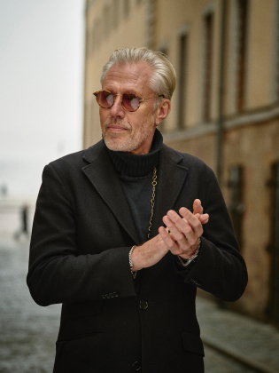 Carl-Johan Vallgren står på en stadsgata klädd i svart kappa och solglasögon, med en lugn och självsäker blick.