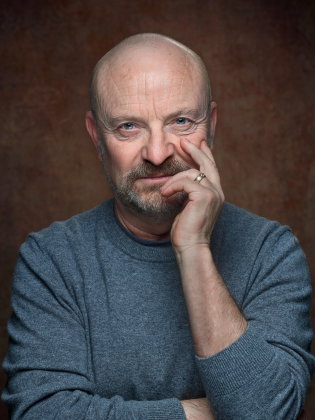 Jonas Gardell sitter framför en mörk bakgrund med handen mot ansiktet i ett intensivt och personligt porträtt.