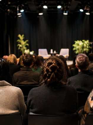 Publik sitter vänd mot en upplyst scen med två fåtöljer och gröna växter inför ett samtal eller scenframträdande.