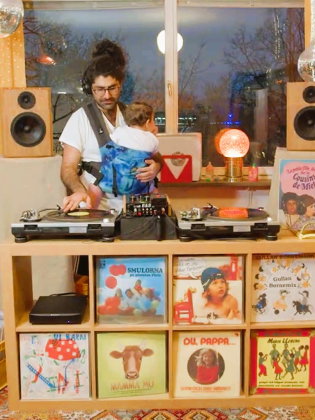 Person står vid skivspelare med ett barn i bärsele i ett vardagsrum fyllt med vinylskivor och högtalare.