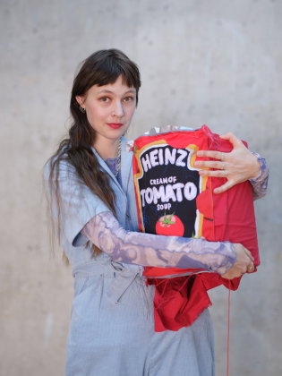 En person håller ett rött konstobjekt som liknar en Heinz-tomatsoppsförpackning framför en betongvägg.
