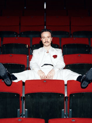 Man i vit kostym sitter ensam bland röda teaterstolar med benen utsträckta över stolsraderna och blickar mot kameran.