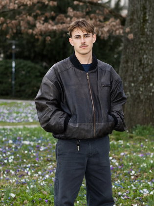 Nelio Biedermann står i ett blommande grönområde i ett avslappnat helkroppsporträtt som kombinerar ungdomlig energi med stillhet.
